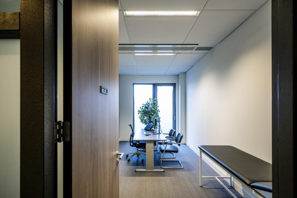 Modern ingerichte dokterspraktijk aan de Laan van de Helende Meesters met bureau, stoelen, onderzoekstafel en grote raampartij.